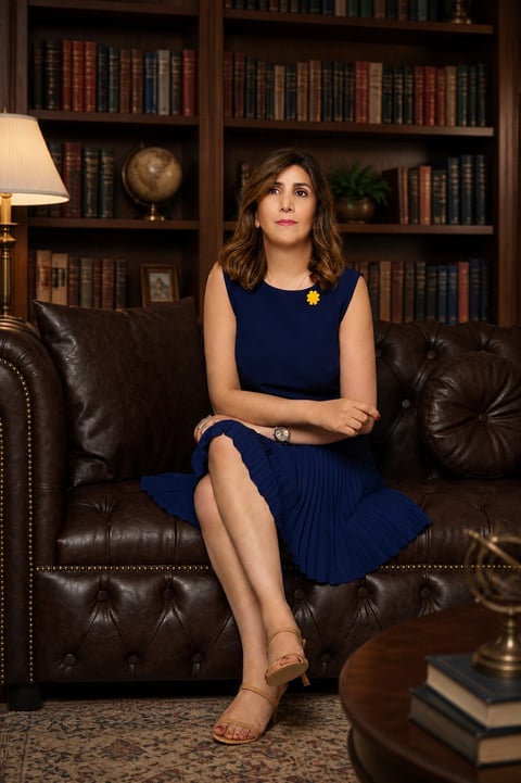 Woman in navy blue dress sitting on leather sofa in front of bookshelf with lamp and globe