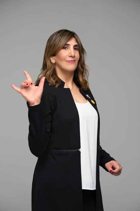 Professional woman in black blazer and white top making a peace sign gesture against a gray background